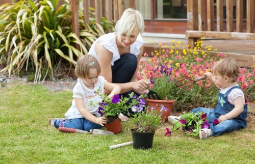 A woman with light blonde hair, wearing a white top, is sitting on a well-maintained green lawn in a garden, tending to a variety of flowering plants in pots and on the ground. She is accompanied by two young children, both with light brown hair, one on each side, engaged in planting or watering the flowers. The garden features a lush grass area with neat borders containing colorful flowering plants, including pink and purple blooms, and leafy shrubs, creating a vibrant outdoor space. In the background, a wooden deck or patio extends from a house with large glass doors, surrounded by tall ornamental grasses and other lush greenery. The scene is illuminated by natural sunlight, suggesting a bright and clear day, emphasizing the well-kept appearance of the garden and outdoor space, which could be part of a residential property in the Bow area, suitable for gardening and outdoor maintenance services.