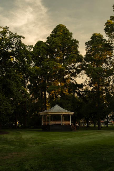 A landscaped park area featuring a small, octagonal pavilion with a light grey, shingle-style roof and wooden railings, situated on a neatly maintained grassy lawn. Tall, mature trees with dense green