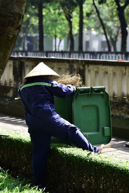 A gardener dressed in a navy uniform with reflective stripes and wearing a traditional conical straw hat is outdoors, leaning into a green wheelie bin with one leg raised on a neatly trimmed hedge. Th